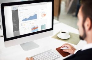Homem de terno sentado em frente a um computador de mesa, observando uma tela com gráficos e relatórios de desempenho. Há uma xícara de café ao lado do teclado. Homem realizando monitoramento de site, analisando dados e gráficos no computador.