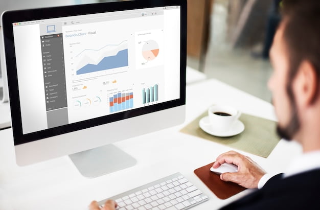 Homem de terno sentado em frente a um computador de mesa, observando uma tela com gráficos e relatórios de desempenho. Há uma xícara de café ao lado do teclado. Homem realizando monitoramento de site, analisando dados e gráficos no computador.