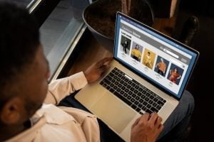 A imagem mostra um close-up de um homem fazendo compras com um laptop, ilustrando o processo de como criar sites WordPress. O homem está sentado à mesa, digitando no teclado do laptop. Ao lado do laptop, há um cartão de crédito, sugerindo que ele está realizando uma compra online. O fundo da imagem é desfocado, destacando o foco na ação de uso do laptop. A cena transmite uma atmosfera de e-commerce e atividade digital. Como criar sites wordpress