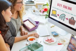 Duas mulheres sentadas em frente a um computador, sorrindo enquanto olham para a tela que exibe a frase "Buy Online". Elas estão cercadas por itens de papelaria e uma xícara de chá. Duas mulheres discutem sobre como criar minha loja virtual, com foco em compras online.