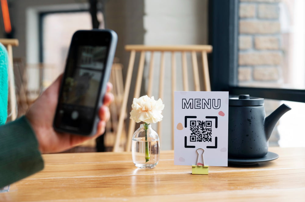 Um cliente em um café está usando seu smartphone para escanear um QR code em um pequeno cartaz que diz "Menu" sobre uma mesa de madeira. O QR code direciona para o menu digital do café, exemplificando o uso prático de QR codes em restaurantes.